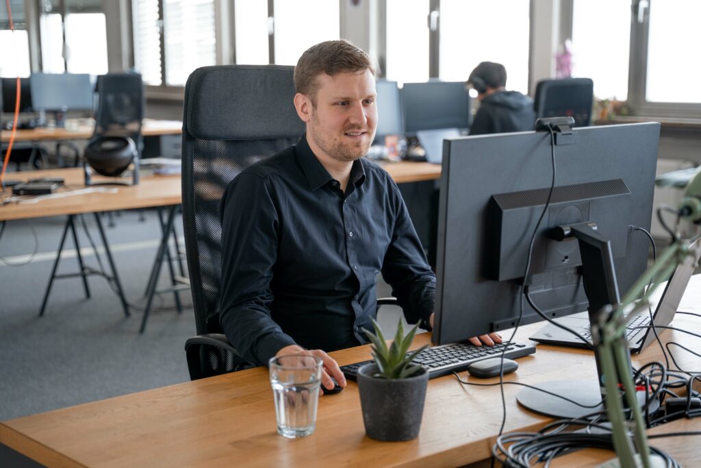 Axel is wearing a black shirt and sitting in front of a large computer monitor. He has a small plant and a glass of water on his right side. In the slightly blurry background, another employer can be seen wearing headphones and focused at work.