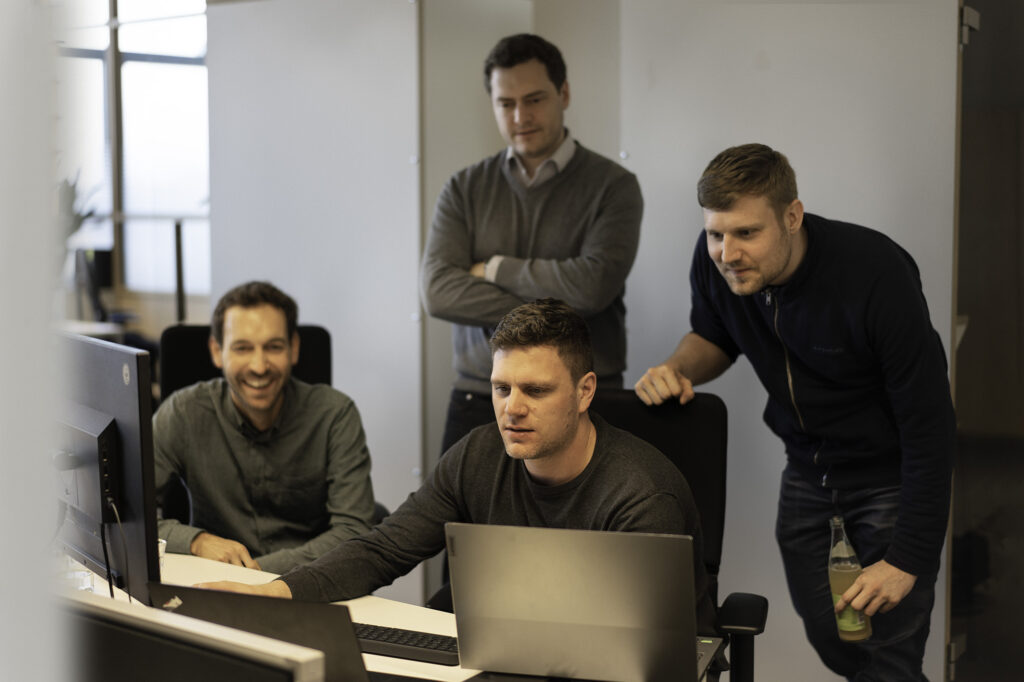 Four men are staring at a monitor and the laptop. The left of the image is slightly blurred. Two men are sitting, both wearing grey shirts. The men on the right has a smile on his face. Behind them, two men are standing. The one on the right has his arms crossed and a grey sweater. On his left, the other man has a dark blue sweater on and is approaching the screen with a curious expression.